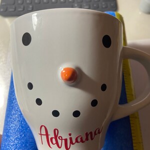 May include: White ceramic mug with a snowman design. The snowman has black eyes, a red nose, and black buttons. The mug has the name "Adriana" written in red cursive.