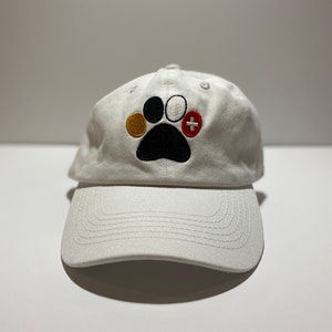 May include: White baseball cap with a black paw print embroidered on the front. The paw print has three colored circles inside: gold, white, and red with a plus sign.