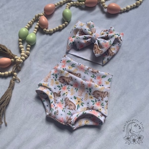 Könnte beinhalten: Weiße Baby-Blümchenhose mit einem Blumenmuster mit Hasen und Schmetterlingen. Ein passendes weißes Stirnband mit einer großen Schleife ist ebenfalls enthalten.
