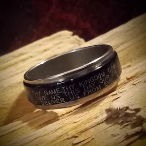 May include: A silver ring with a black band featuring engraved text. The text reads "THY NAME. THY KINGDOM COME GIVE US THIS DAY OUR DA DELIVER US FROM EVIL..." The ring is set against a wooden surface.