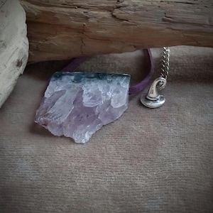 Könnte beinhalten: Ein Anhänger aus rohem Amethystkristall mit einem silberfarbenen Hexenhut-Anhänger. Der lila Kristall ist an einem lila Band befestigt. Der Anhänger wird auf einer braunen Oberfläche mit einem hölzernen Hintergrund präsentiert.