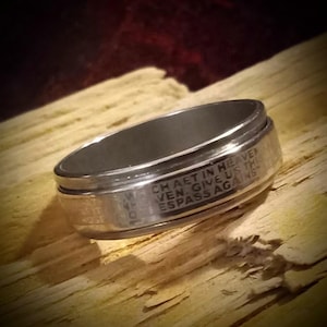May include: A silver-colored ring with engraved text. The ring has a dark interior and a polished exterior. The text includes the words "CHAET IN HEAVEN" and "GIVE US THIS". The ring is set against a wooden background.