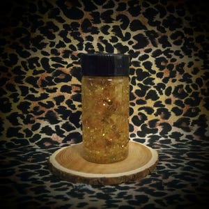 Puede incluir: Un pequeño frasco de vidrio con tapa negra lleno de purpurina dorada. El frasco está sentado sobre una rodaja de madera.