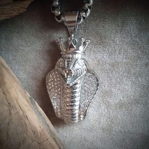 Könnte beinhalten: Silberfarbener Anhänger mit einer detaillierten Kobra-Schlangenform mit einer Krone auf dem Kopf. Die Schlange ist mit klaren Strasssteinen verziert.