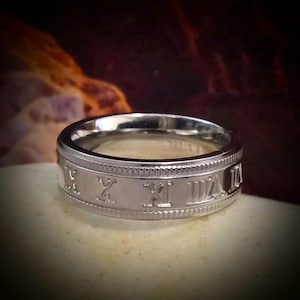 May include: A silver band ring with Roman numeral markings around the band.