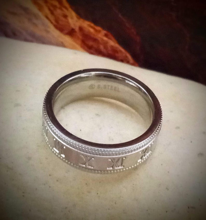 Peut inclure: Une bague en argent avec des chiffres romains grav&eacute;s sur le bord. La bague est en acier inoxydable.