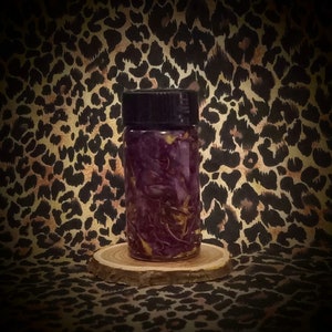 May include: A small, clear glass bottle with a black cap, filled with purple and yellow botanicals. The bottle sits on a small, round wooden coaster. The background features a leopard print pattern in shades of brown and black.