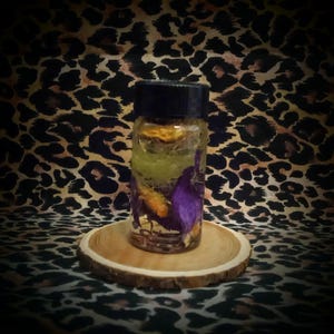 May include: A small glass bottle with a black cap, filled with colorful dried flowers and herbs. The bottle sits on a round wooden coaster. The background features a leopard print pattern.