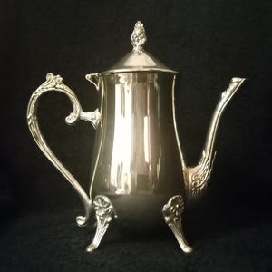 May include: A silver-colored coffee pot with a lid and a curved handle. The pot has a spout and sits on three decorative feet. The handle and spout have ornate detailing. The lid is topped with a decorative finial.