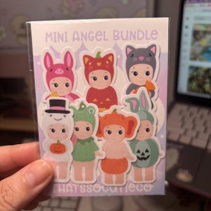 May include: A Mini Angel Bundle sticker sheet featuring seven cartoon figures in various costumes, including a pig, pumpkin, cat, ghost with a top hat, frog, orange fruit, and a green rabbit. The backing card has light purple and white colours.