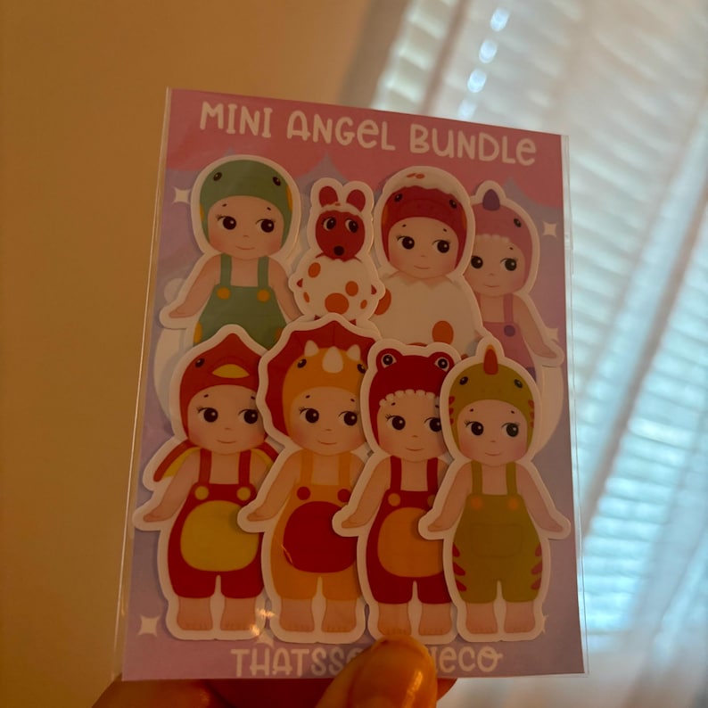 May include: A Mini Angel Bundle sticker sheet featuring seven cartoon figures in various animal-themed costumes, including a green frog, a red dinosaur, and a yellow triceratops. The background is light purple with white stars and the text Mini Angel Bundle.