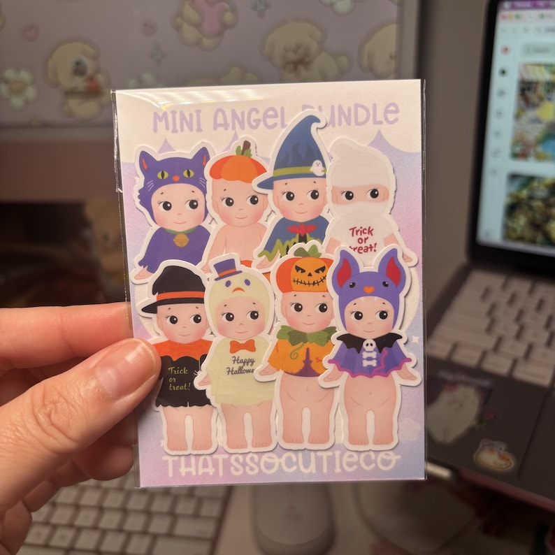 May include: A sticker sheet titled MINI ANGEL BUNDLE featuring eight cartoon baby figures in various Halloween costumes, including a cat, pumpkin, witch, ghost, bat, and snowman. The background is a light purple gradient with the text THATSSOCUTIECO.