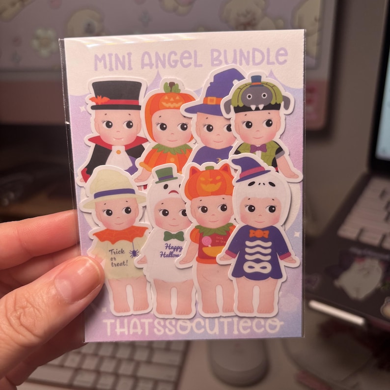 May include: A Mini Angel Bundle sticker pack featuring eight cartoon baby figures in various Halloween costumes, including a pumpkin, witch, and skeleton. The backing card displays the text Mini Angel Bundle and thatssocutieco in purple.