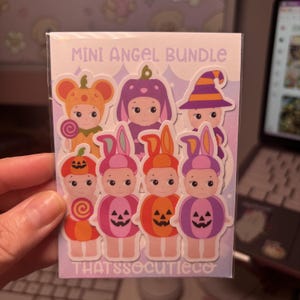 May include: A Mini Angel Bundle sticker sheet featuring seven cartoon characters in Halloween costumes, including pumpkin and witch outfits. The characters are colourful with orange, purple, and pink tones. The text reads MINI ANGEL BUNDLE and THATSSOCUTIECO.