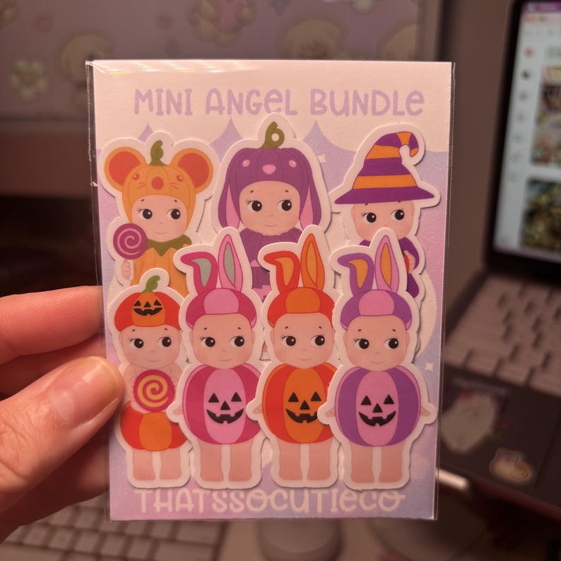 May include: A Mini Angel Bundle sticker sheet featuring seven cartoon characters in Halloween costumes, including pumpkin and witch outfits. The characters are colourful with orange, purple, and pink tones. The text reads MINI ANGEL BUNDLE and THATSSOCUTIECO.