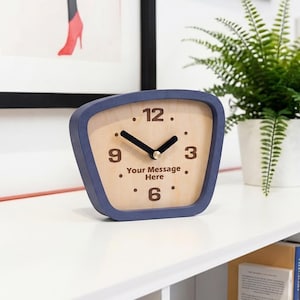 May include: A wooden clock with a blue frame displays the time. The clock face is light-colored with black numbers and hands. The text "Your Message Here" is printed below the 9. A red lamp and a potted fern are also visible.
