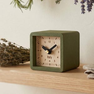May include: A square olive green analog clock with a light beige face, black hour and minute hands, and the word Italy printed in black. The clock features a simple numerical dial with markings for each hour, housed in a matte green frame.