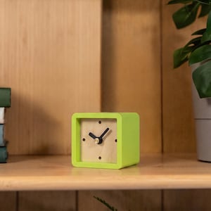 May include: A square-shaped analog clock with a bright lime green frame and a light wood-toned face. The clock features four dark circular hour markers at the twelve, three, six, and nine o'clock positions, along with two black hands and a gold center.