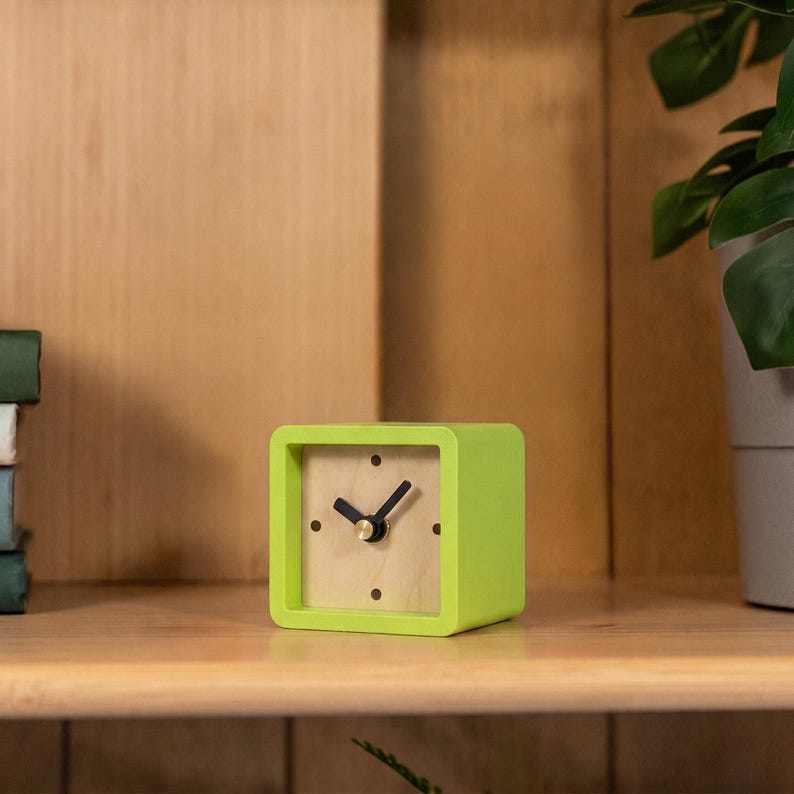 May include: A square-shaped analog clock with a bright lime green frame and a light wood-toned face. The clock features four dark circular hour markers at the twelve, three, six, and nine o'clock positions, along with two black hands and a gold center.
