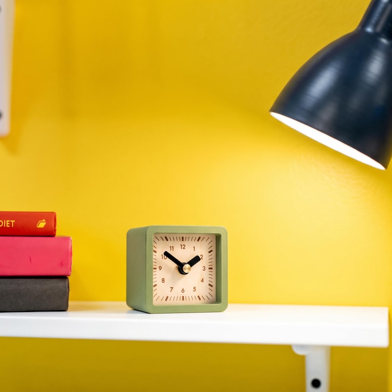 May include: A square-shaped analog alarm clock with a sage green frame and a light-colored face. The clock features black hour and minute hands, black numerals from 1 to 12, and black tick marks for each minute around the perimeter of the dial.