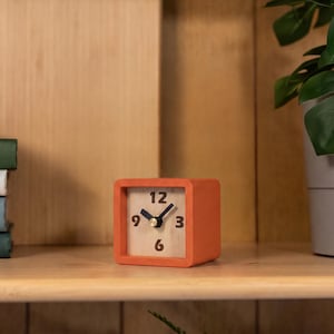 May include: A square analog clock with a matte orange frame and a light wood grain face. The clock features dark brown numerals at the 12, 3, 6, and 9 positions, along with two dark brown hands and a small gold-toned center cap for the movement.