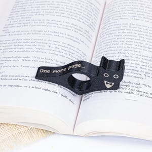 May include: A black cat-shaped page holder with a gold glitter finish. The holder has a ring to hold a page and the text "One more page..." is printed on the holder.