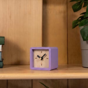 May include: A square-shaped analog clock with a light purple frame and a light-colored wooden face. The clock features two black hands and a small gold-colored center pin. The face has simple black tick marks for the hour indicators.