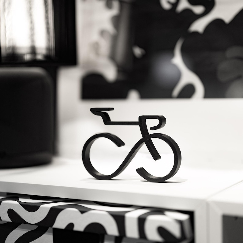Puede incluir: Escultura de bicicleta de metal negro con un dise&ntilde;o minimalista. La escultura est&aacute; colocada sobre una superficie blanca, con una caja con estampado en blanco y negro en primer plano. El fondo presenta una l&aacute;mpara negra y arte abstracto.