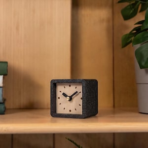 May include: A square desk clock with a dark gray speckled concrete frame and a light wood face. The clock face features black hour and minute hands, a gold center pin, and small circular icons representing moon phases instead of numbers.