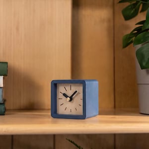 May include: A square-shaped analog desk clock with a matte blue frame and a light wood-toned face. The clock features simple black hour markers, two black hands, and a small gold-colored center pin. The design is compact and geometric.