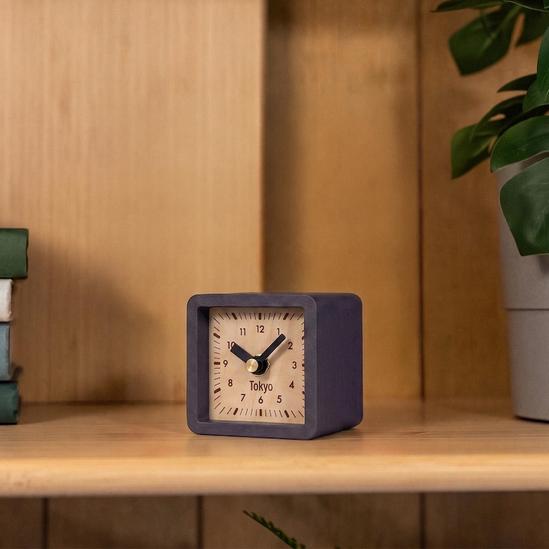 May include: A square-shaped analog clock with a dark purple frame and a light wood-toned face. The clock features black hour and minute hands, a gold-colored center pin, black numerical markings from 1 to 12, and the word Tokyo printed in black.