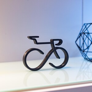 Puede incluir: Una escultura de bicicleta de metal negro con un dise&ntilde;o minimalista. La escultura presenta l&iacute;neas curvas que forman las ruedas y el cuadro, con un manillar recto. La bicicleta se exhibe sobre una superficie blanca.