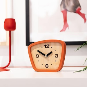 May include: A modern, orange-framed wooden clock with a light wood face. Black hands point to the time, with black numbers at 3, 6, 9, and 12. The clock sits on a white surface, with a red candle holder to the left.