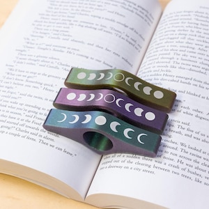 May include: Set of three iridescent bookmark clips with moon phases. Each clip is a different color: green, purple, and blue. The moon phases are white and are printed on the clips.