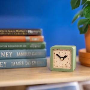 May include: A small, square, sage green analog alarm clock with a light-colored face, black numerals, and thick black hands. The clock features a central gold-toned pin and is positioned on a light wood surface next to a stack of books.