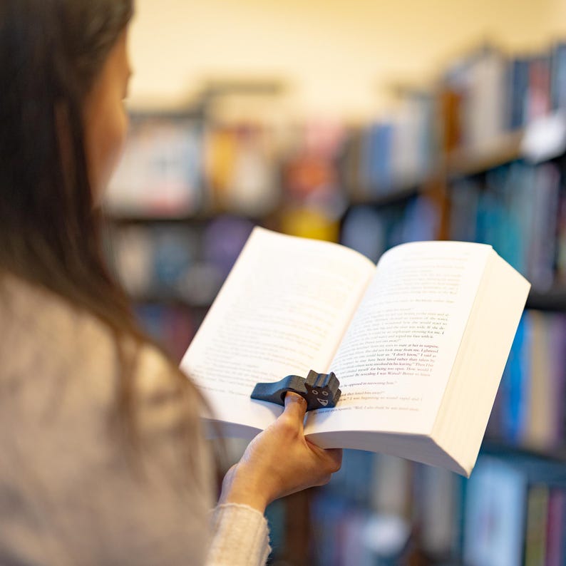 May include: A person is reading a book with a black cat-shaped page holder. The book is open and the page holder is holding the page in place. The person is sitting in a library with bookshelves in the background.