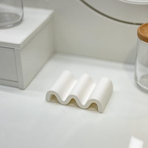 Może przedstawiać: Biała ceramiczna mydelniczka o falistym wzorze. Mydelniczka stoi na białej powierzchni.