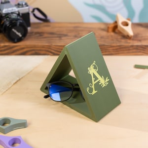 May include: A green triangular glasses holder with a gold letter "A" and floral design.  A pair of blue-tinted glasses rests in the holder.