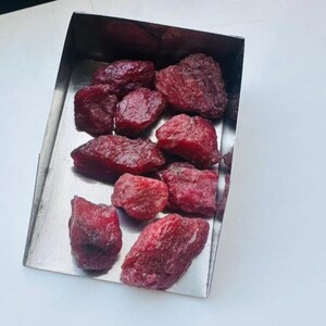 May include: A selection of raw ruby gemstones displayed in a stainless steel tray. The rubies are a deep red hue, with a slightly uneven texture. The tray is rectangular and has a polished, metallic finish.