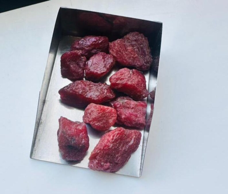 May include: A selection of raw ruby gemstones displayed in a stainless steel tray. The rubies are a deep red hue, with a slightly uneven texture. The tray is rectangular and has a polished, metallic finish.