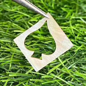 May include: A square, iridescent, mother-of-pearl spade shape is held by tweezers against a backdrop of green grass. The spade shape has a cutout design, and the surface reflects light with a pearlescent sheen.