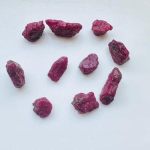 May include: A selection of raw ruby gemstones in various shapes and sizes. The deep red stones are scattered on a white surface, highlighting their natural, unpolished state. These gemstones are suitable for jewellery making or collecting. The stones are measured in grams.