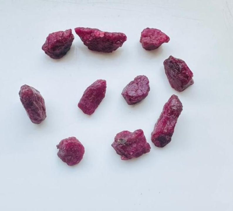 May include: A selection of raw ruby gemstones in various shapes and sizes. The deep red stones are scattered on a white surface, highlighting their natural, unpolished state. These gemstones are suitable for jewellery making or collecting. The stones are measured in grams.