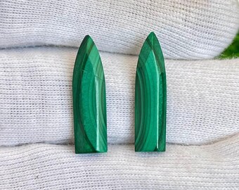 May include: Two polished malachite gemstones with a vibrant green color and distinctive banded patterns. The gemstones have a pointed, elongated shape, and are displayed on a white fabric background.