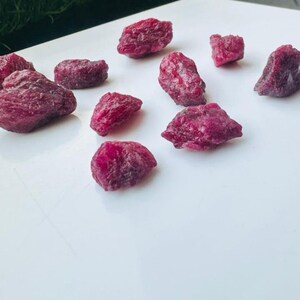 May include: A selection of raw ruby gemstones, displaying a deep red hue. The gemstones, of various sizes and shapes, are presented on a white surface. The lighting accentuates the natural texture and colour of the rubies, showcasing their raw beauty.