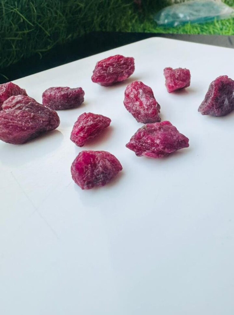 May include: A selection of raw ruby gemstones, displaying a deep red hue. The gemstones, of various sizes and shapes, are presented on a white surface. The lighting accentuates the natural texture and colour of the rubies, showcasing their raw beauty.