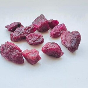 May include: A cluster of raw ruby gemstones, each with a deep red colour and a rough, textured surface. The uncut rubies are scattered on a white background, highlighting their natural, irregular forms. These gemstones are suitable for jewelry making.