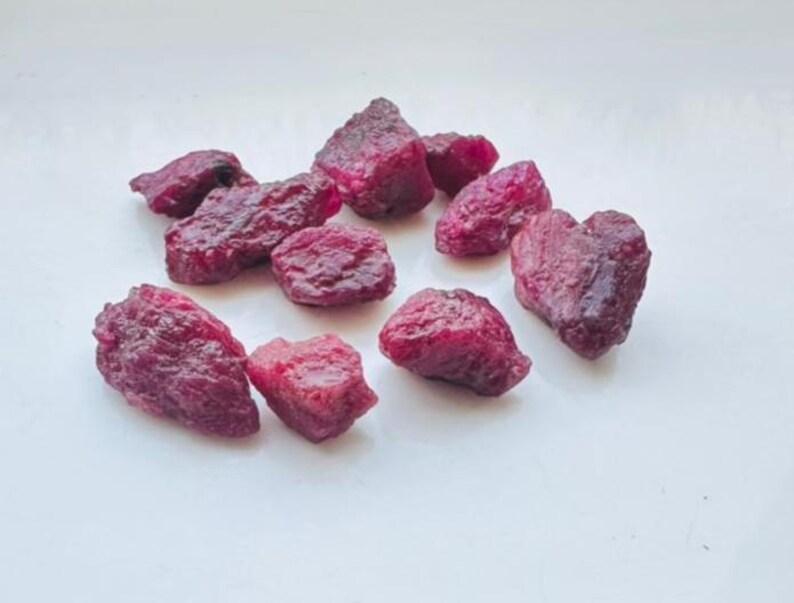 May include: A cluster of raw ruby gemstones, each with a deep red colour and a rough, textured surface. The uncut rubies are scattered on a white background, highlighting their natural, irregular forms. These gemstones are suitable for jewelry making.