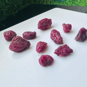 May include: A selection of rough ruby gemstones in various sizes. The raw rubies exhibit a deep red colour and are arranged on a white surface. The gemstones have a natural, unpolished appearance, displaying their organic textures and shapes.