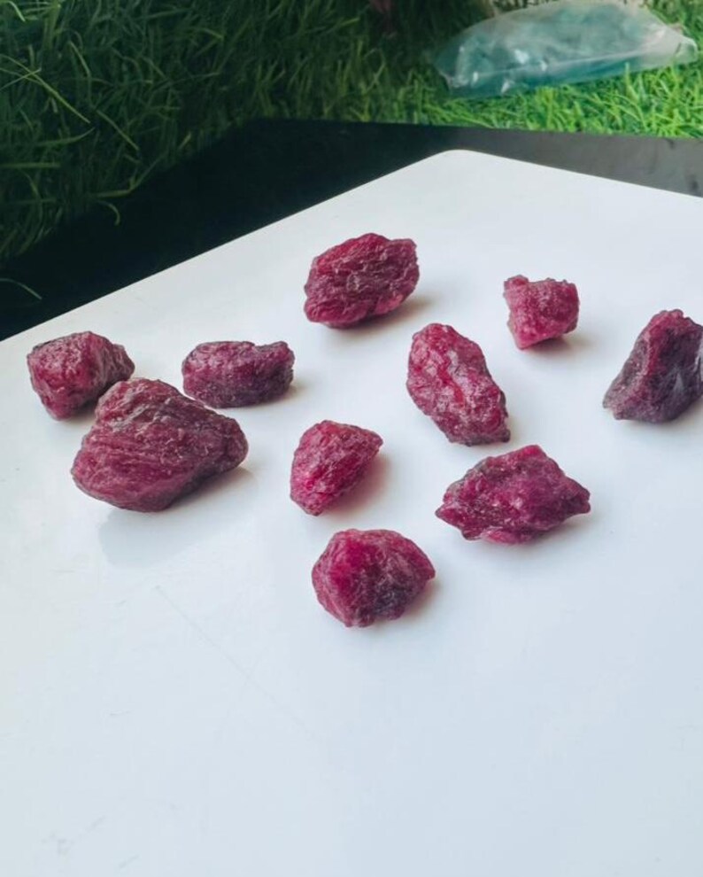 May include: A selection of rough ruby gemstones in various sizes. The raw rubies exhibit a deep red colour and are arranged on a white surface. The gemstones have a natural, unpolished appearance, displaying their organic textures and shapes.